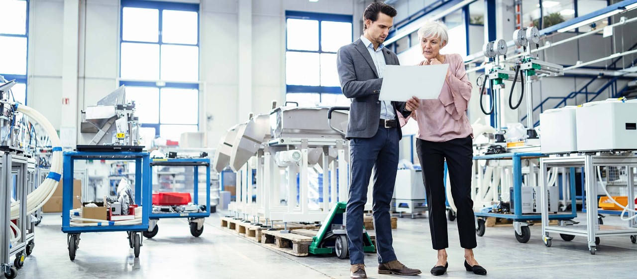 Ein Mann und eine Frau schauen auf Unterlage in einer Produktionshalle